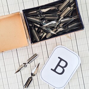 K&ouml;nnte beinhalten: Eine Sammlung metallisch silberner Kalligrafiefedern in einer rechteckigen blauen Pappschachtel, mit zwei weiteren Federn au&szlig;erhalb der Schachtel neben einer wei&szlig;en rechteckigen Karte, auf der der schwarze Buchstabe B abgedruckt ist.