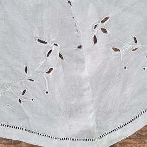 Puede incluir: Un tejido de lino blanco con delicados bordados calados en un patr&oacute;n de hojas y enredaderas. La tela tiene una textura tejida visible y un dobladillo acabado con un detalle de punto de escalera a lo largo del borde inferior.