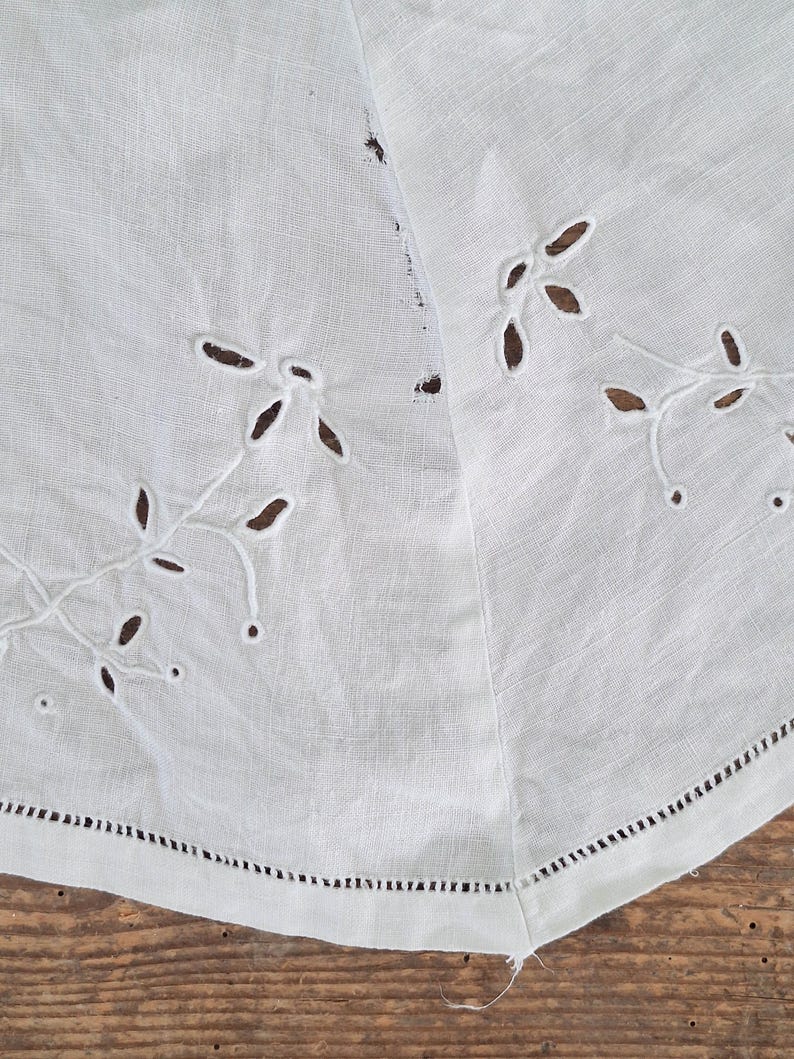 Puede incluir: Un tejido de lino blanco con delicados bordados calados en un patr&oacute;n de hojas y enredaderas. La tela tiene una textura tejida visible y un dobladillo acabado con un detalle de punto de escalera a lo largo del borde inferior.