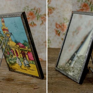 May include: Two antique picture frames with metal stands. One frame displays a colorful image of a building with figures, the other a reflective surface. The frames have a dark, ornate border and are set on a wooden surface.
