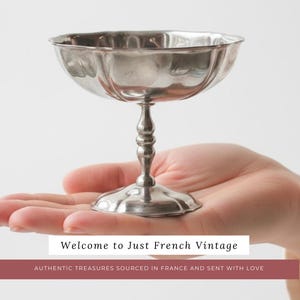 May include: A silver-colored, stemmed dessert cup with a scalloped rim is held in a person's hand. The cup has a wide, shallow bowl and a decorative, baluster-shaped stem. The text "Welcome to Just French Vintage" is displayed.