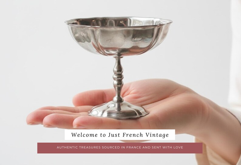 May include: A silver-colored, stemmed dessert cup with a scalloped rim is held in a person's hand. The cup has a wide, shallow bowl and a decorative, baluster-shaped stem. The text "Welcome to Just French Vintage" is displayed.
