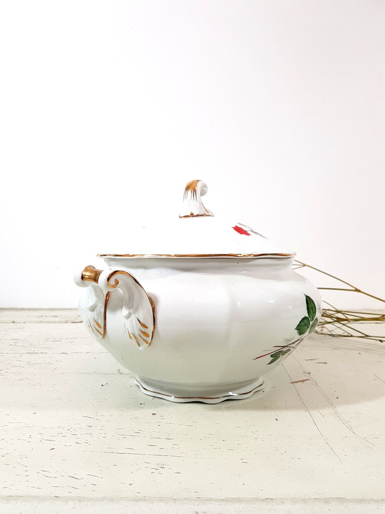 May include: White porcelain soup tureen with a gold trim and floral design. The tureen has a lid with a gold knob and a handle on each side.