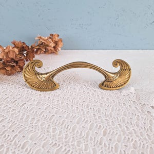 May include: A brass-toned metal drawer pull featuring a curved handle and two symmetrical, fan-shaped ends with intricate engraved detailing. The hardware has a weathered, antique finish with visible dark patina in the recessed areas.