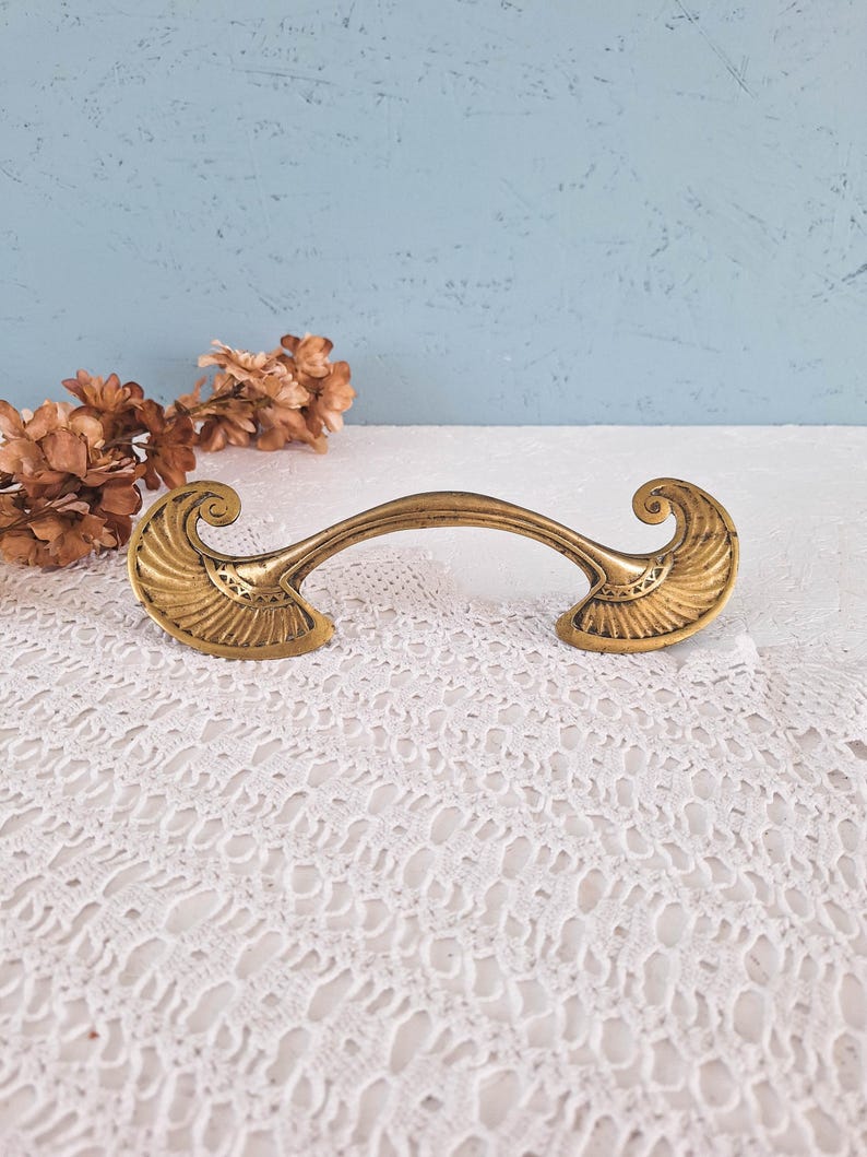 May include: A brass-toned metal drawer pull featuring a curved handle and two symmetrical, fan-shaped ends with intricate engraved detailing. The hardware has a weathered, antique finish with visible dark patina in the recessed areas.