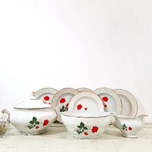 May include: A white porcelain dinnerware set with a red rose pattern. The set includes a soup tureen, a serving bowl, a creamer, and four dinner plates.