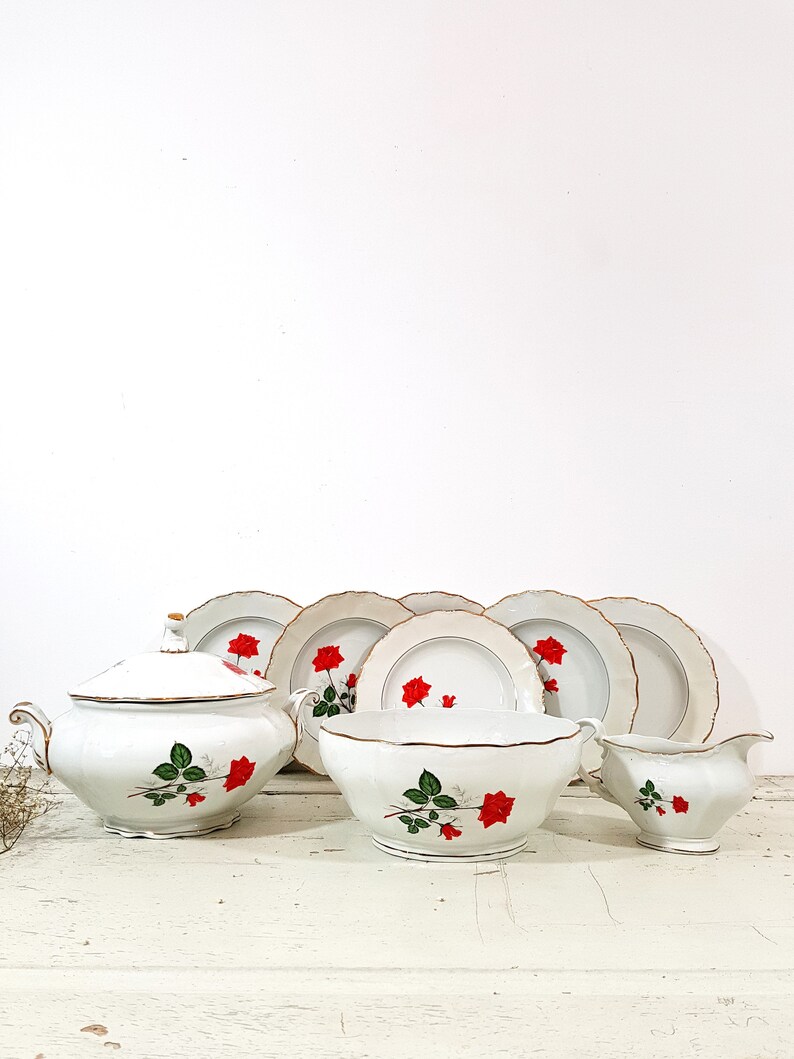 May include: A white porcelain dinnerware set with a red rose pattern. The set includes a soup tureen, a serving bowl, a creamer, and four dinner plates.