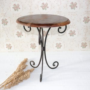 Puede incluir: Una mesa redonda de madera con una base de metal negro con un diseño decorativo de desplazamiento. La mesa está sobre una superficie blanca.