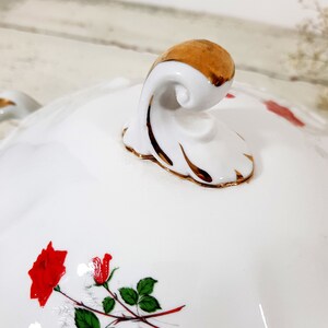 May include: White porcelain serving dish with a gold trim and a floral design featuring red roses. The dish has a decorative handle with a gold accent.
