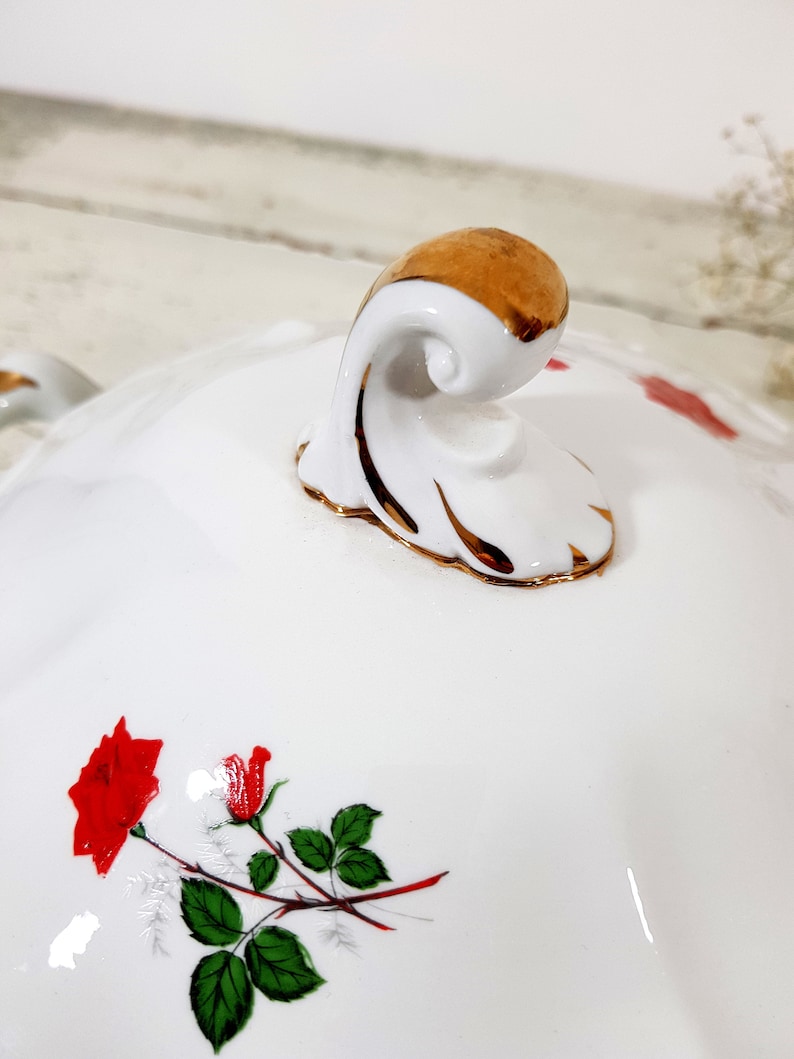 May include: White porcelain serving dish with a gold trim and a floral design featuring red roses. The dish has a decorative handle with a gold accent.