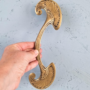 May include: A gold-toned metal door handle featuring an elongated, curved central bar that connects two symmetrical, fan-shaped ends. Each end displays a textured, radiating pattern with a small spiral detail at the base of the curve.