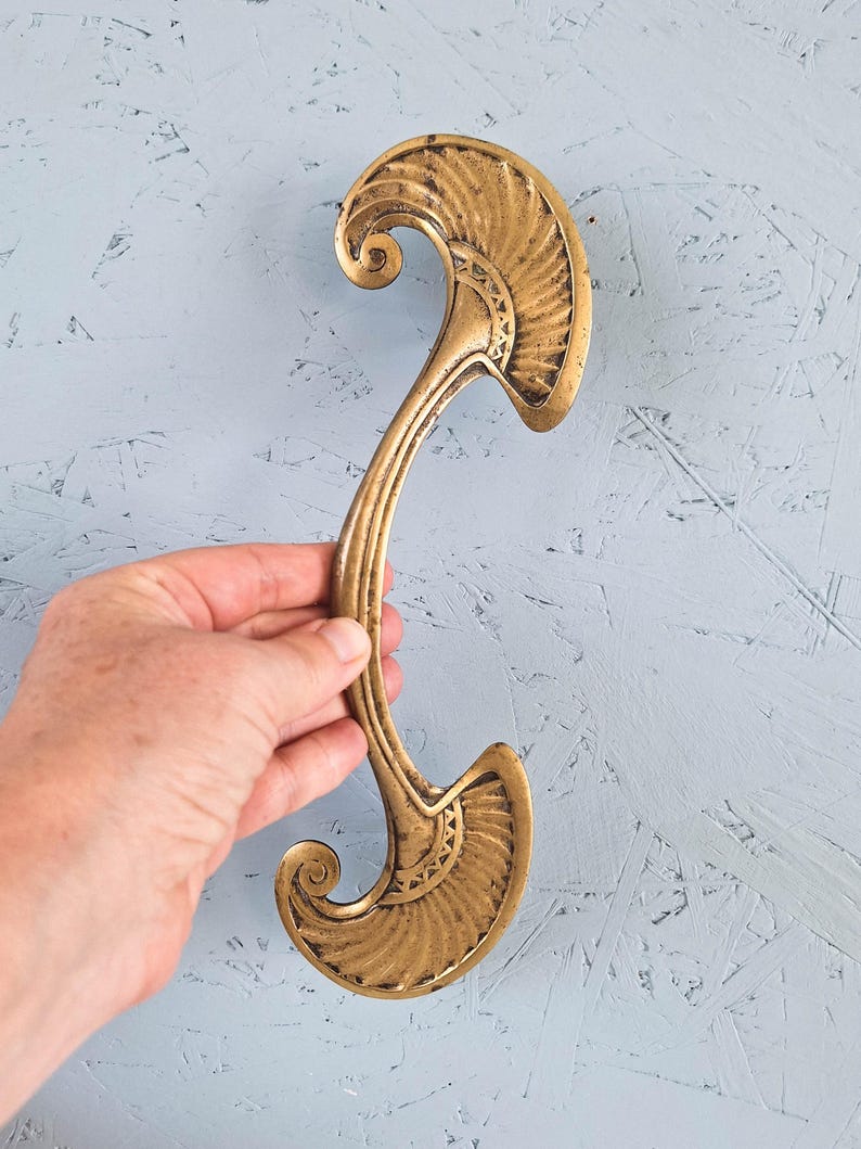 May include: A long, curved brass door handle featuring symmetrical, fan-shaped ends with intricate, engraved geometric patterns. The metal has a weathered, antique finish with visible dark patina in the recessed areas of the decorative design.