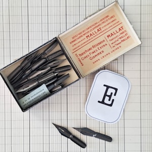 K&ouml;nnte beinhalten: Eine Vintage-Schachtel von Mallat mit dunklen Kalligrafiefedern aus Metall, dazu lose Federn und ein wei&szlig;es rechteckiges Etikett mit einem schwarzen Buchstaben E. Die Innenseite des Deckels zeigt roten und schwarzen Text in franz&ouml;sischer Sprache.