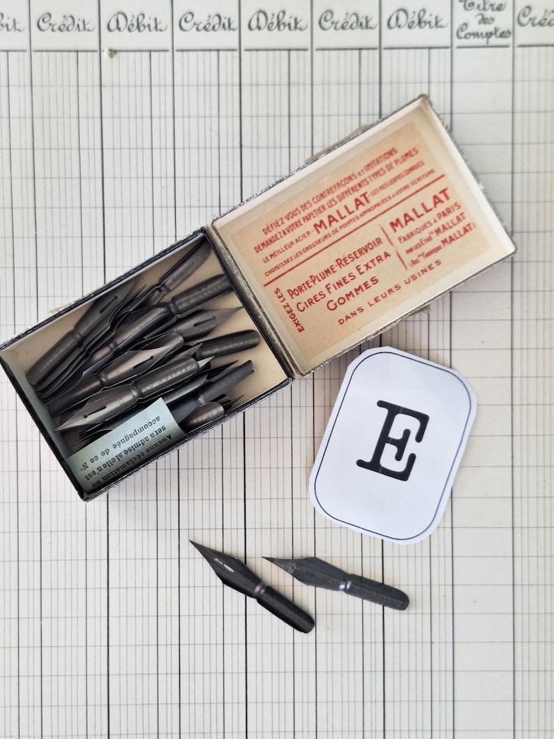 K&ouml;nnte beinhalten: Eine Vintage-Schachtel von Mallat mit dunklen Kalligrafiefedern aus Metall, dazu lose Federn und ein wei&szlig;es rechteckiges Etikett mit einem schwarzen Buchstaben E. Die Innenseite des Deckels zeigt roten und schwarzen Text in franz&ouml;sischer Sprache.