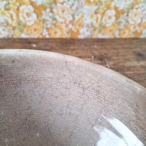 May include: Close-up of a vintage, off-white ceramic bowl with a cracked, aged appearance. The bowl's surface is covered in a network of fine cracks, suggesting age and use. The background features a floral wallpaper and a wooden surface.