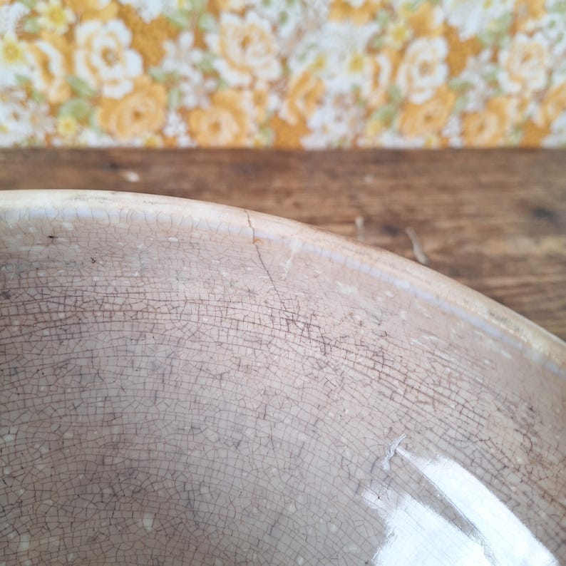 May include: Close-up of a vintage, off-white ceramic bowl with a cracked, aged appearance. The bowl's surface is covered in a network of fine cracks, suggesting age and use. The background features a floral wallpaper and a wooden surface.