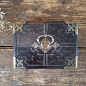Puede incluir: Una caja rectangular de madera oscura con incrustaciones metálicas plateadas de motivos florales, cuatro protectores de esquina de latón decorativos y un asa central de metal plateado con una placa circular y detalles de filigrana.