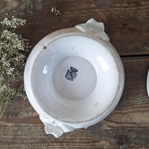May include: A white ceramic bowl with a floral design on the rim. The bowl has a light brown rim and a black and white emblem on the bottom. The bowl is on a rustic wooden surface.