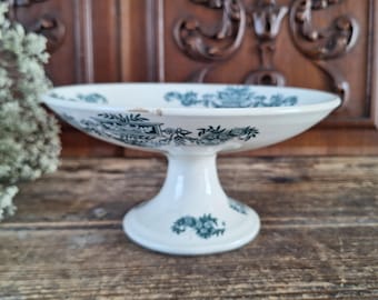 May include: A white ceramic pedestal bowl featuring a dark green transferware pattern of floral motifs and architectural urns. The bowl has a wide, shallow basin supported by a sturdy, flared base, with a small chip visible on the rim.