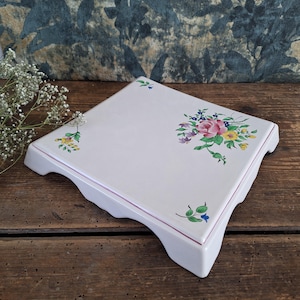 May include: A square white ceramic stand with a glossy finish, featuring a painted floral motif with a central pink rose, yellow and blue flowers, and green leaves. The stand has four short, curved legs and a thin pink line along the edge.