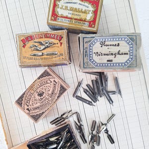 K&ouml;nnte beinhalten: Eine Sammlung von vier alten Pappschachteln mit Metallfedern f&uuml;r die Kalligrafie. Die Schachteln sind mit kunstvoller Typografie und Illustrationen versehen, darunter Texte wie 100 PLUMES METALLIQUES, LA RENOMM&Eacute;E, Plumes Birmingham und PLUME GAULOISE, sowie lose silberfarbene Schreibfedern.