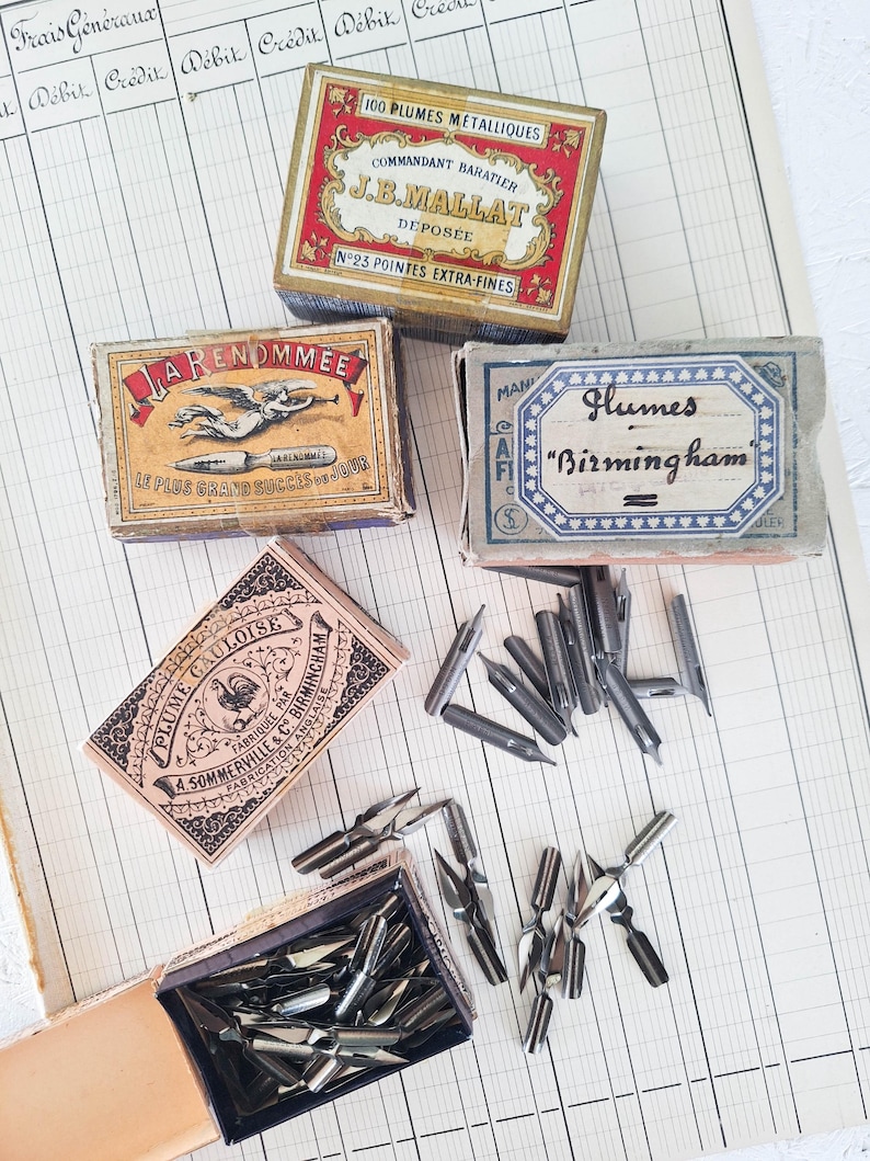 K&ouml;nnte beinhalten: Eine Sammlung von vier alten Pappschachteln mit Metallfedern f&uuml;r die Kalligrafie. Die Schachteln sind mit kunstvoller Typografie und Illustrationen versehen, darunter Texte wie 100 PLUMES METALLIQUES, LA RENOMM&Eacute;E, Plumes Birmingham und PLUME GAULOISE, sowie lose silberfarbene Schreibfedern.
