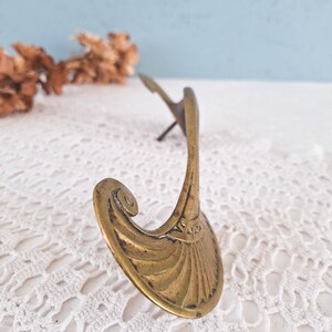 May include: A brass wall hook featuring a curved, fan-shaped base with engraved linear detailing and a prominent scroll accent at the top. The metal surface displays a weathered patina, and a single mounting pin extends from the back of the hook.