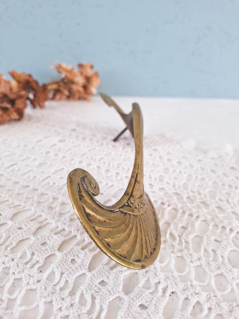May include: A brass wall hook featuring a curved, fan-shaped base with engraved linear detailing and a prominent scroll accent at the top. The metal surface displays a weathered patina, and a single mounting pin extends from the back of the hook.
