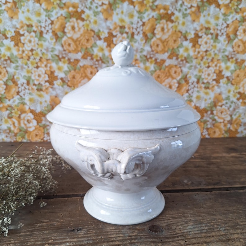 May include: A white ceramic lidded serving dish with a pedestal base. The dish features a decorative floral design on the lid and handles. The background is a floral wallpaper.