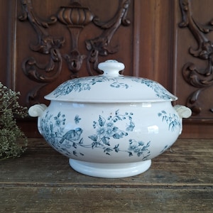 Puede incluir: Fuente de cerámica blanca con tapa, adornada con motivos florales y pájaros azules. La fuente tiene dos asas laterales y una base redonda. La tapa está rematada con un pequeño pomo. Fondo de madera oscura.