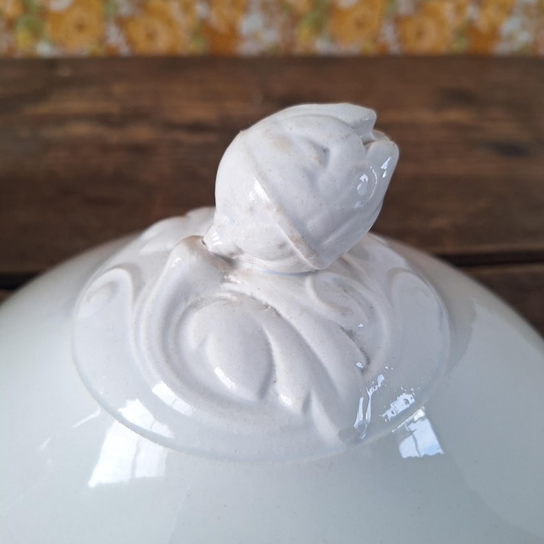 May include: A white ceramic serving dish lid with a tulip-shaped handle. The handle and the rim of the lid feature embossed floral designs. The lid has a glossy finish and is likely part of a serving set.