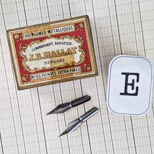 K&ouml;nnte beinhalten: Eine rechteckige Pappschachtel mit roten und goldenen Zierr&auml;ndern, beschriftet mit 100 PLUMES METALLIQUES COMMANDANT BARATIER J.B.MALLAT DEPOSEE N&deg;23 POINTES EXTRA-FINES, zwei Metallfedern und ein wei&szlig;es Etikett mit einem schwarzen E.