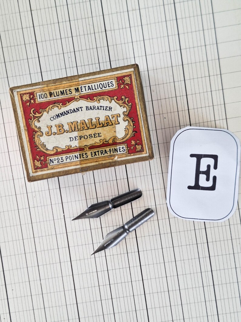 K&ouml;nnte beinhalten: Eine rechteckige Pappschachtel mit roten und goldenen Zierr&auml;ndern, beschriftet mit 100 PLUMES METALLIQUES COMMANDANT BARATIER J.B.MALLAT DEPOSEE N&deg;23 POINTES EXTRA-FINES, zwei Metallfedern und ein wei&szlig;es Etikett mit einem schwarzen E.