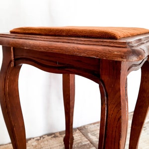 Peut inclure: Petit tabouret en bois antique avec un dessus carr&eacute; et des pieds incurv&eacute;s. Le tabouret est teint&eacute; d'une couleur brun chaud et le si&egrave;ge est recouvert d'un tissu textur&eacute; brun-orang&eacute;. Le design rappelle les meubles vintage.