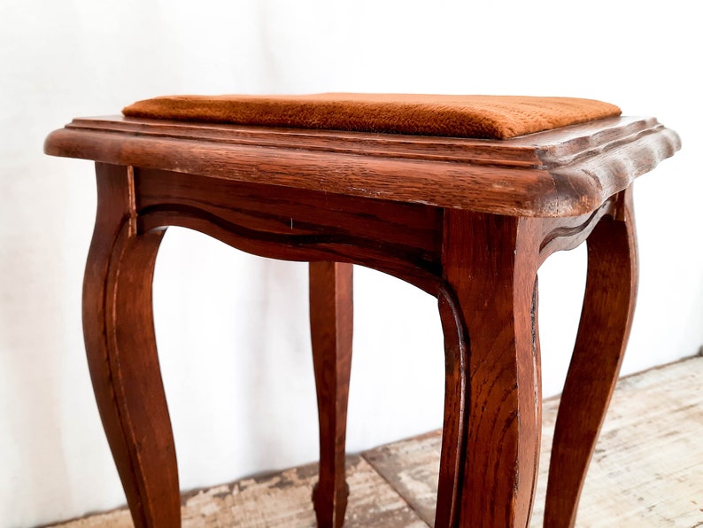 Peut inclure: Petit tabouret en bois antique avec un dessus carr&eacute; et des pieds incurv&eacute;s. Le tabouret est teint&eacute; d'une couleur brun chaud et le si&egrave;ge est recouvert d'un tissu textur&eacute; brun-orang&eacute;. Le design rappelle les meubles vintage.