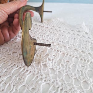 May include: Two curved brass wall hooks with a weathered patina and visible oxidation. Each hook features a flat, rounded mounting plate and a threaded metal screw extending from the back for installation, showing a metallic gold and green finish.