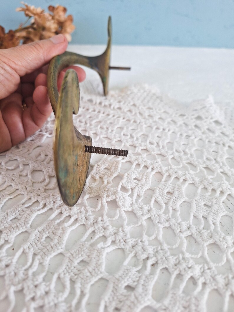 May include: Two curved brass wall hooks with a weathered patina and visible oxidation. Each hook features a flat, rounded mounting plate and a threaded metal screw extending from the back for installation, showing a metallic gold and green finish.