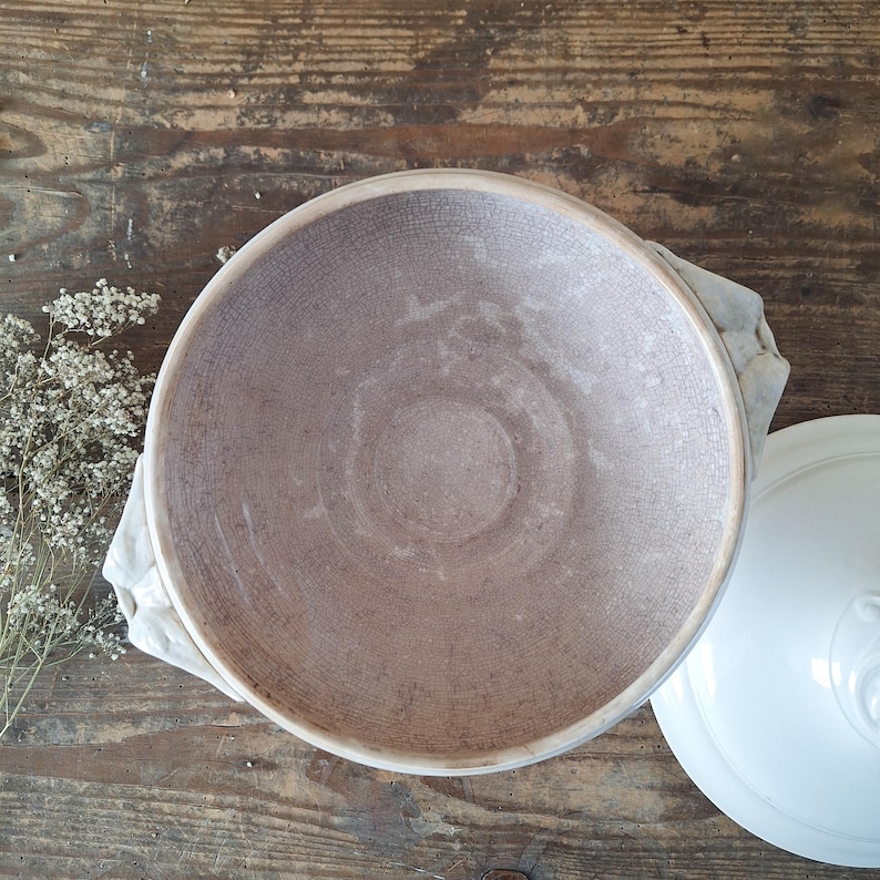 May include: A large, round, antique-style ceramic bowl with a light brown interior and a textured surface. The bowl sits on a white cloth, next to a white lid with a decorative handle. The background is a rustic wooden surface.