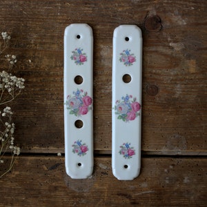 May include: Two white porcelain cabinet pulls with a floral design featuring pink and blue roses. Each pull has three round holes and a hole at each end for mounting. The pulls are displayed on a rustic wooden surface.