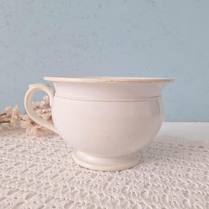 May include: A vintage off-white ceramic chamber pot featuring a rounded basin, a wide flared rim, and a single curved side handle. The piece sits on a circular pedestal base and displays a smooth, glazed finish with subtle signs of age and wear.