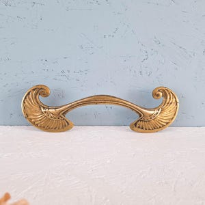 May include: A brass-toned metal drawer pull featuring a curved central bar and two symmetrical, fan-shaped ends with intricate engraved line detailing. The hardware has a weathered, antique finish with visible dark patina in the recessed areas.