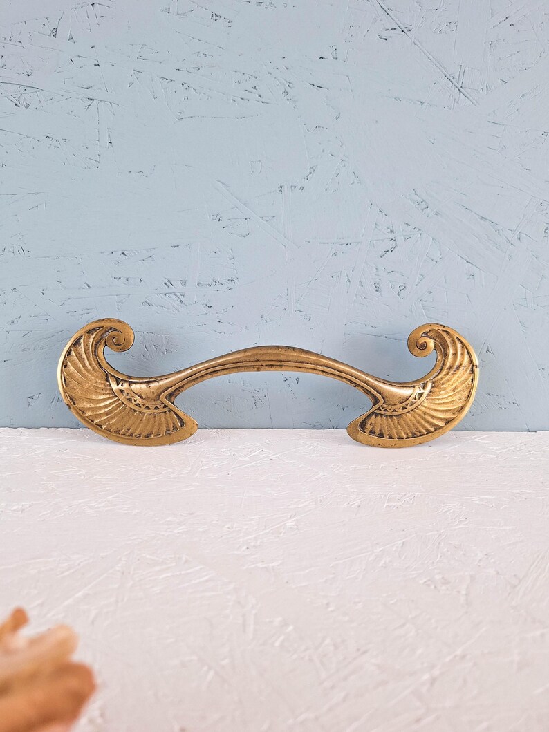 May include: A brass-toned metal drawer pull featuring a curved central bar and two symmetrical, fan-shaped ends with intricate engraved line detailing. The hardware has a weathered, antique finish with visible dark patina in the recessed areas.