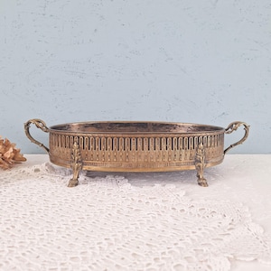 Peut inclure: Un plateau ovale en laiton doté d'une galerie ajourée complexe, de deux poignées latérales ornées et de quatre pieds décoratifs. La surface métallique présente une patine sombre et vieillie avec une texture visible sur le bord et la base.