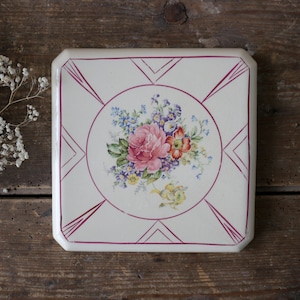 May include: A square ceramic tile with a cream-colored base featuring a central floral bouquet of pink, blue, yellow, and green flowers. The design is framed by a thin magenta circular border and geometric magenta corner accents on the edges.