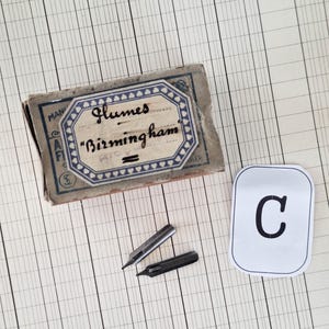 K&ouml;nnte beinhalten: Eine rechteckige Vintage-Pappschachtel mit der Aufschrift Glumes Birmingham, die zwei metallische Schreibfedern enth&auml;lt, eine silberfarbene und eine dunkelgraue, neben einer wei&szlig;en rechteckigen Karte mit dem schwarzen Buchstaben C.