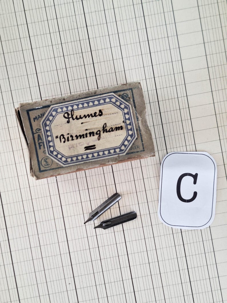 K&ouml;nnte beinhalten: Eine rechteckige Vintage-Pappschachtel mit der Aufschrift Glumes Birmingham, die zwei metallische Schreibfedern enth&auml;lt, eine silberfarbene und eine dunkelgraue, neben einer wei&szlig;en rechteckigen Karte mit dem schwarzen Buchstaben C.