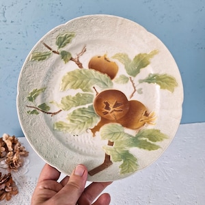 May include: A round ceramic plate featuring a raised, embossed border with a floral pattern. The center displays a hand-painted design of brown medlar fruits on branches with light green leaves against an off-white, textured background.