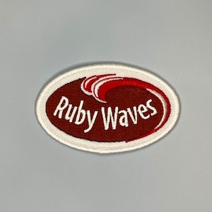 May include: White oval embroidered patch with a red wave and the text "Ruby Waves" in white.