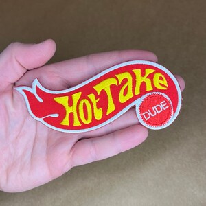 May include: Red and yellow embroidered patch with the text "Hot Take" in a white outline. A smaller red circle with the text "Dude" in white is on the right side of the patch.