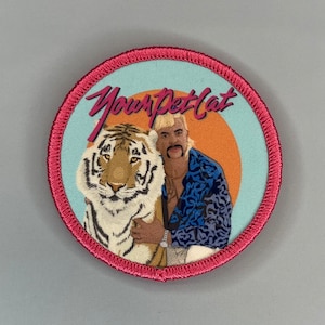 Pode incluir: Um remendo redondo bordado de cor rosa e azul com um tigre e um homem com um topete loiro. O texto "Your Pet Cat" está escrito em letras cor-de-rosa no remendo.